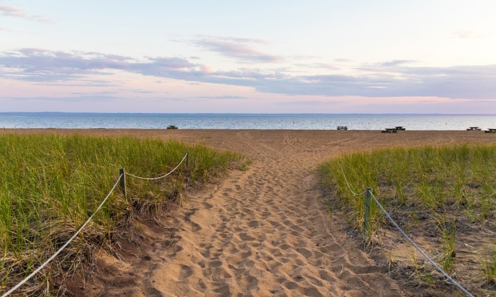 La plage du parc national de la Pointe-Taillon au coucher du soleil. À l’avant-plan, un passage sablonneux et bordé de verdure mène vers la plage. 