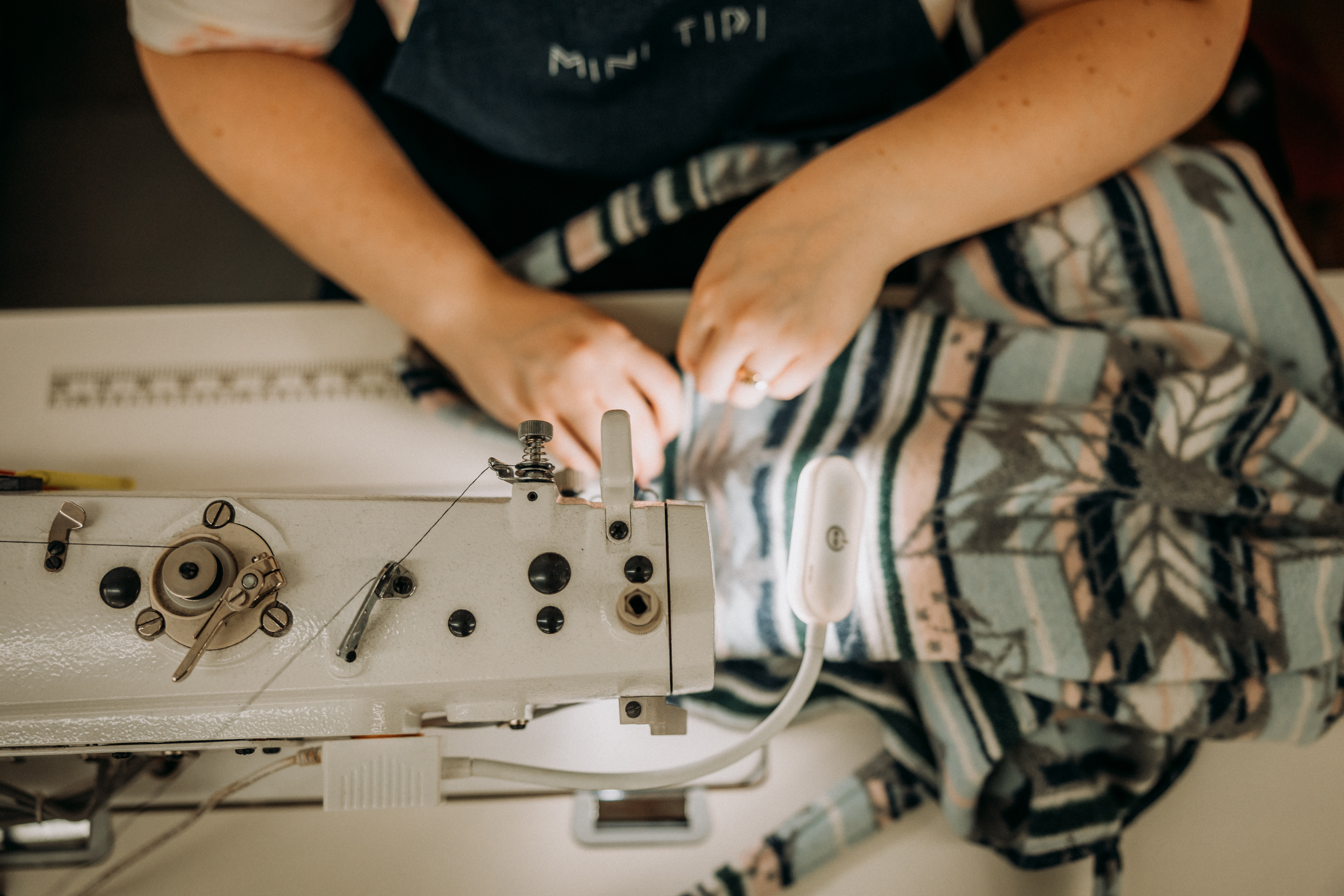 Une vue en gros plan montre les mains d’une travailleuse assise à une machine à coudre, en train de faire la finition d’un produit MINI TIPI orné de motifs traditionnels autochtones.
