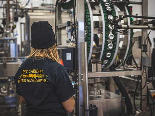 Une employée de la microbrasserie Pit Caribou se tient devant une cuve de brassage, dos à la caméra. Son t-shirt porte la mention “Bière gaspésienne”
