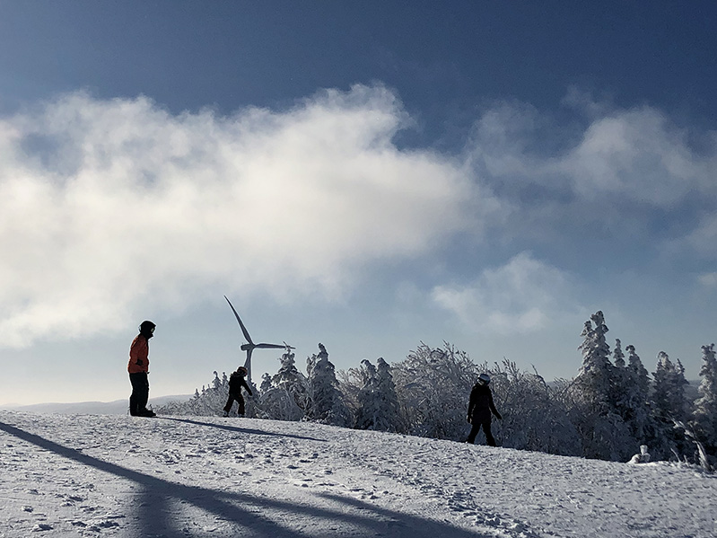 Une famille est tout équipée pour faire de la planche à neige sur une piste enneigée du Mont Miller; une éolienne est visible à l’arrière-plan